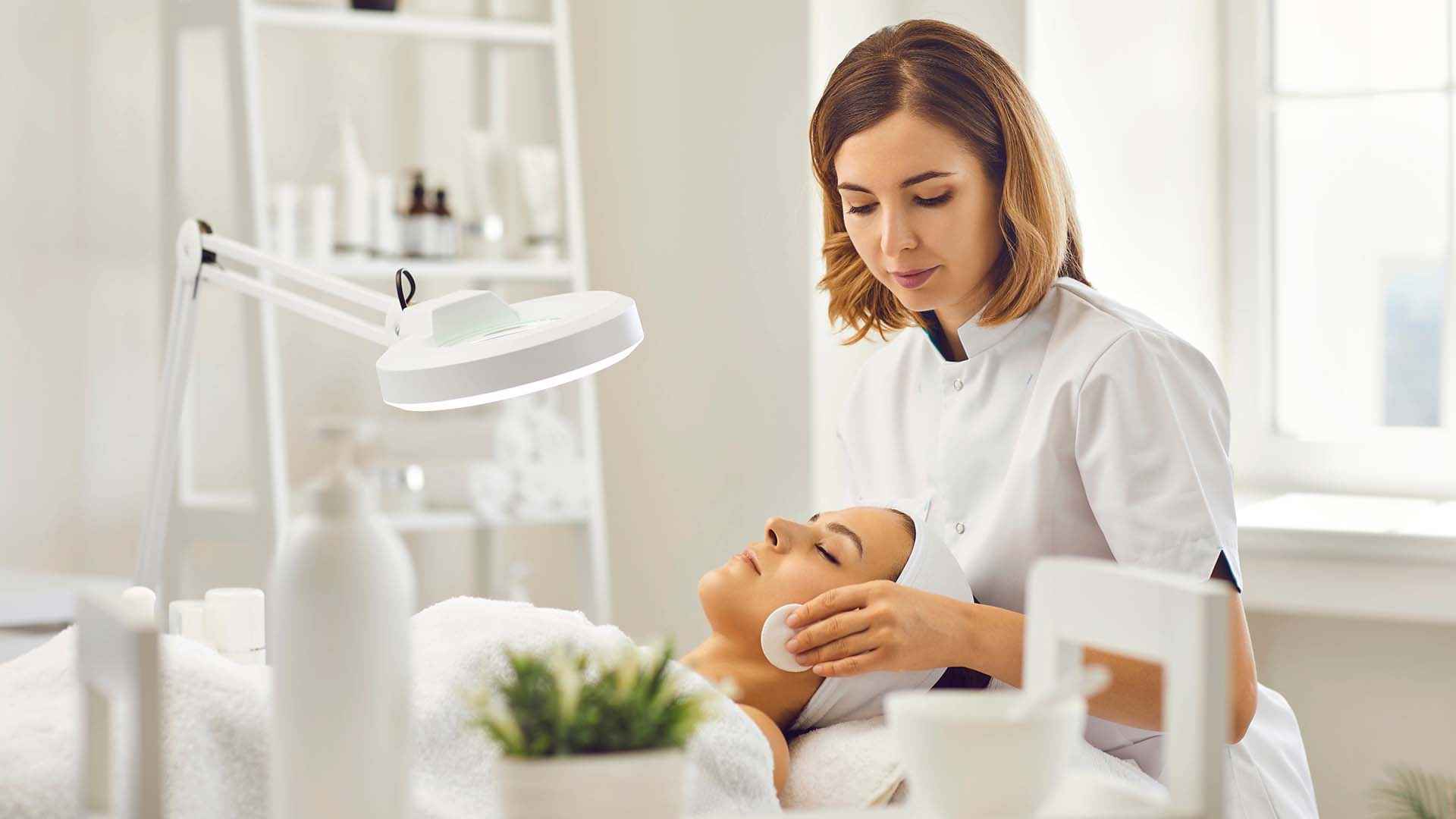 Young cosmetologist or dermatologist cleaning lying womans face with cotton pads in spa salon